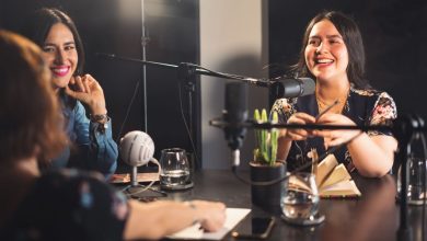 Três mulheres participam de uma gravação descontraída de podcast em um estúdio moderno, diante de microfones.