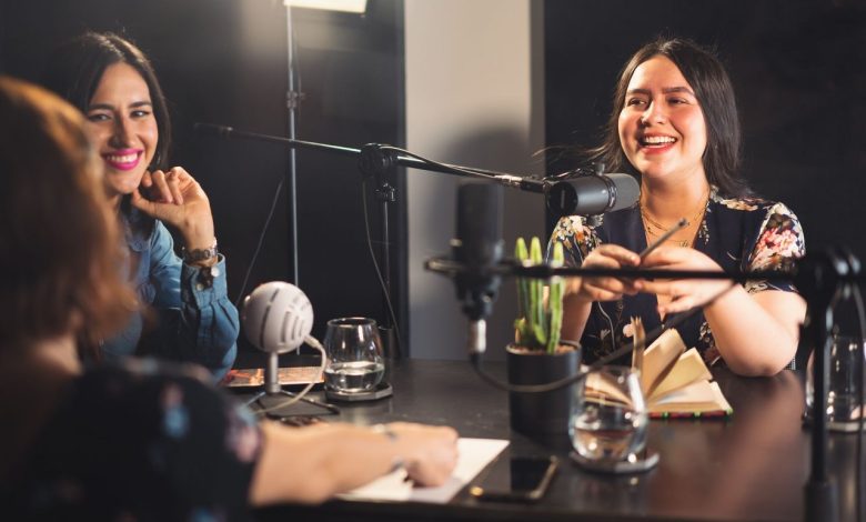 Três mulheres participam de uma gravação descontraída de podcast em um estúdio moderno, diante de microfones.
