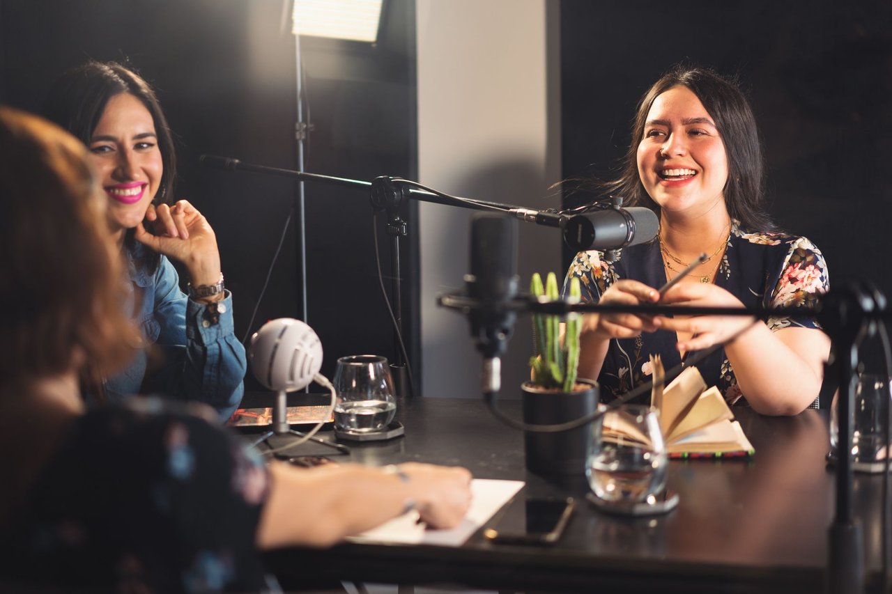 Três mulheres participam de uma gravação descontraída de podcast em um estúdio moderno, diante de microfones.