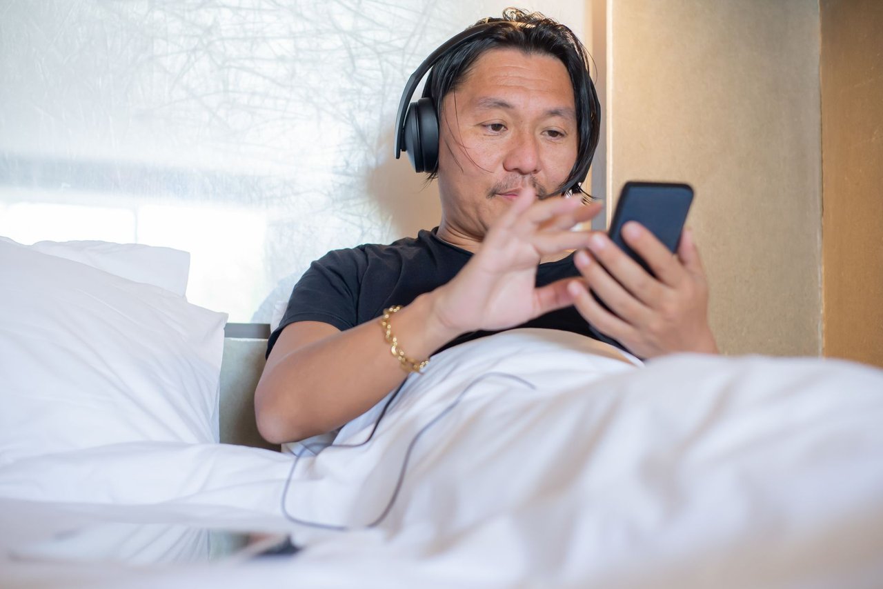Homem asiático usando smartphone e fones de ouvido em um quarto aconchegante.