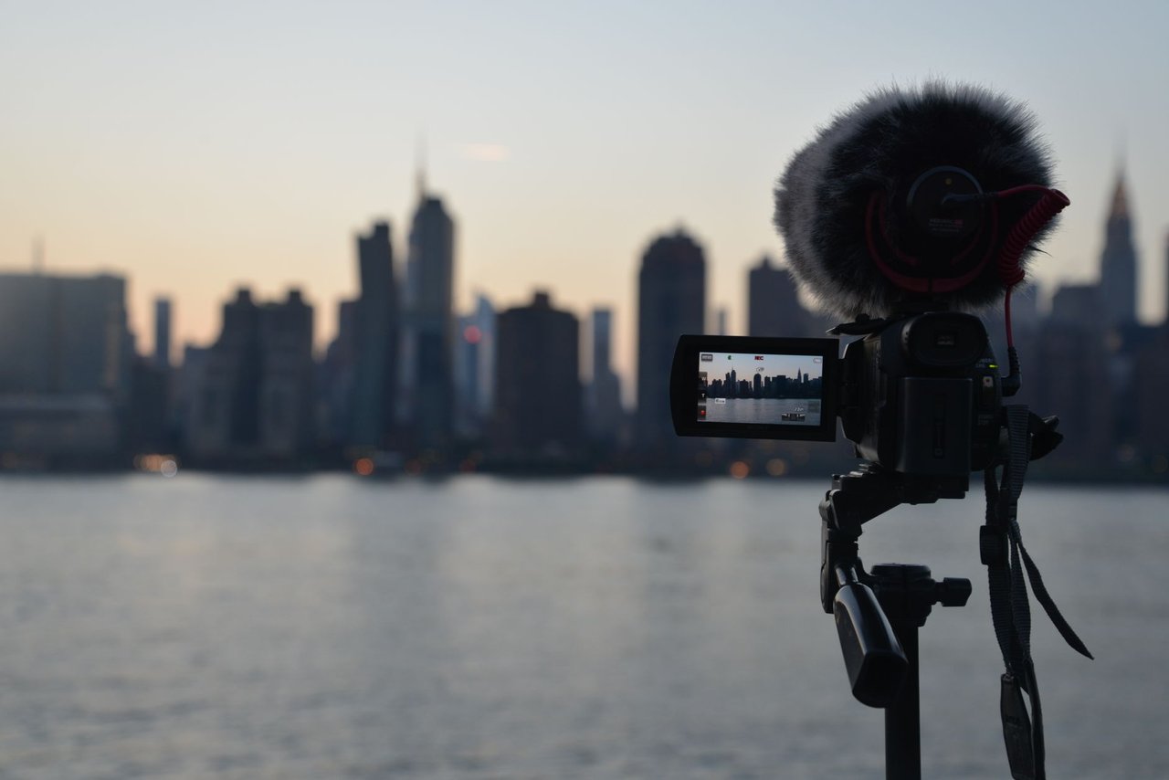 Câmera com microfone diante da paisagem de Nova York ao amanhecer, vista a partir da margem de um rio.